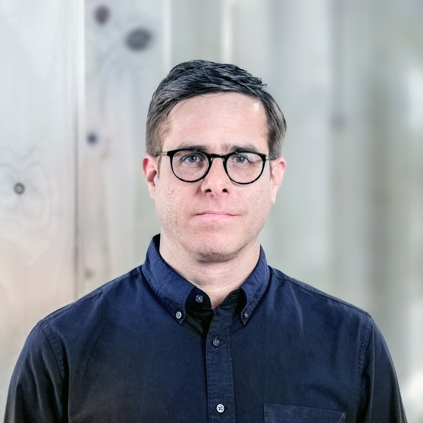 A portrait of a man with short dark hair and round glasses wearing a dark blue button-down shirt against a blurred background.