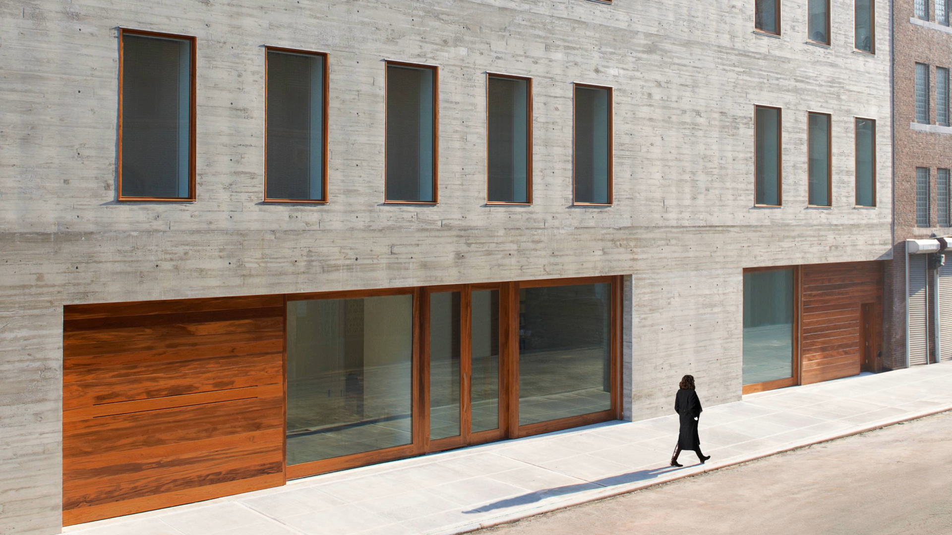 The exterior of the David Zwirner gallery features a multi-story, board-formed concrete facade with rhythmic wooden-framed windows and a person walking along the sidewalk below.