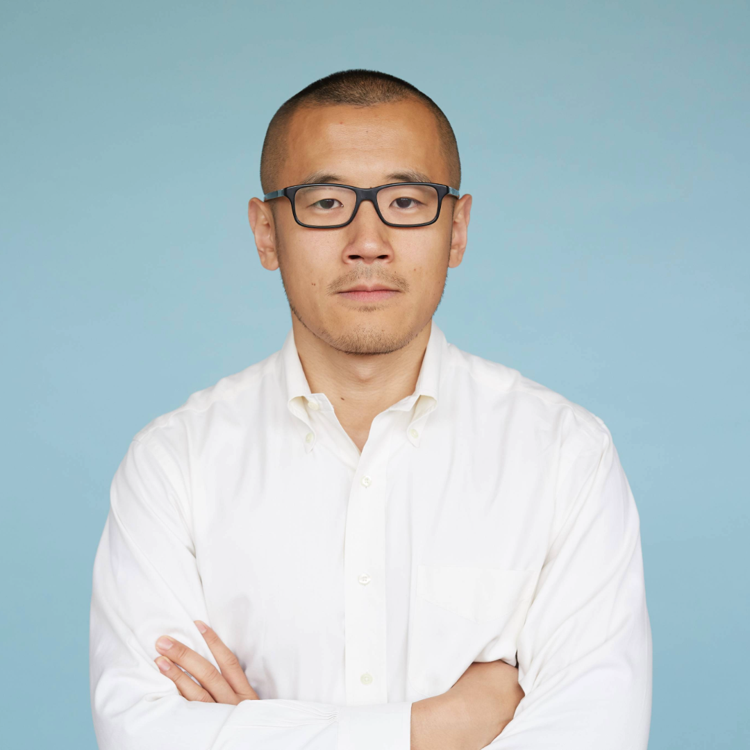 A portrait of a man with short hair and rectangular glasses wearing a white button-down shirt with his arms crossed against a light blue background.