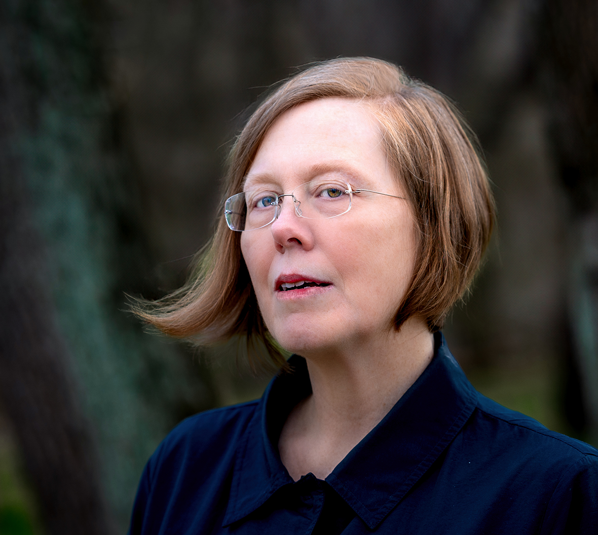 Woman with chin-length strawberry blonde hair wearing a blue jacket in a dark forest