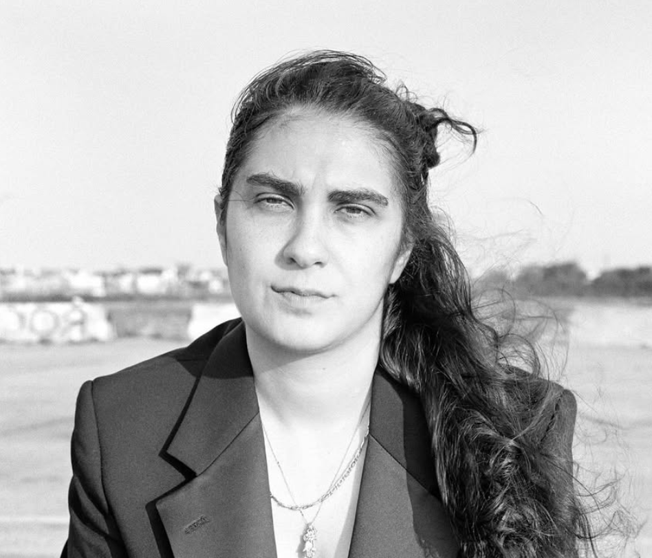 A person with long, dark hair, wearing a blazer jacket and two layered necklaces.