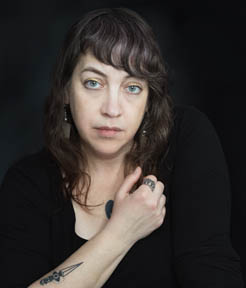 A woman with green eyes and wavy brown hair, styled with asymmetrical bangs.