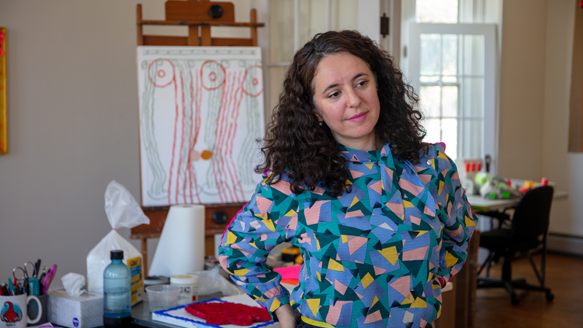 A woman with wavy brown hair and a blue geometric pattern shirt, standing in an art studio.