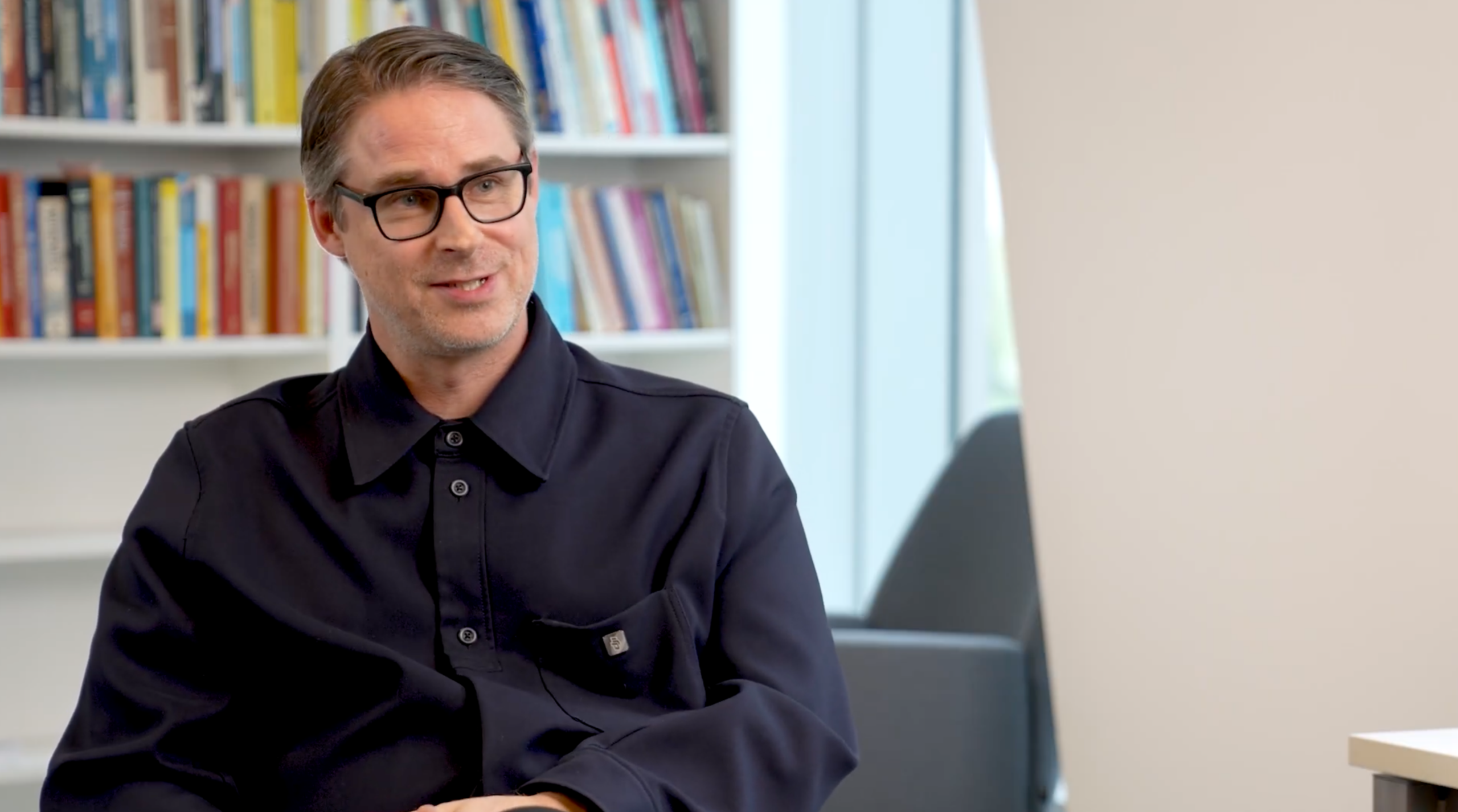 A man wearing a dark blue button down tshirt, sitting in front of a bookcase.