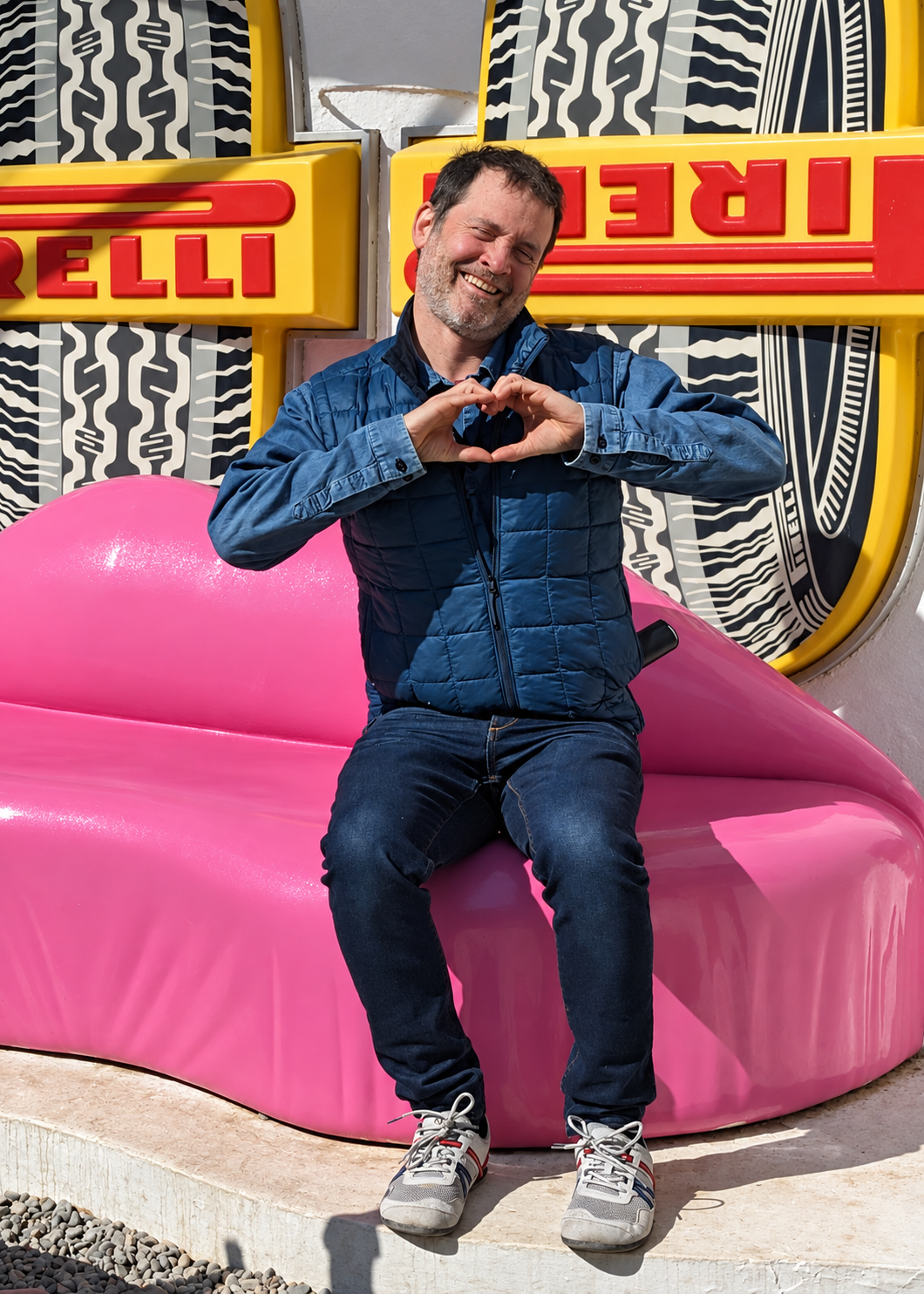 A man sitting on a bright pink couch, making a heart shape with his hands.